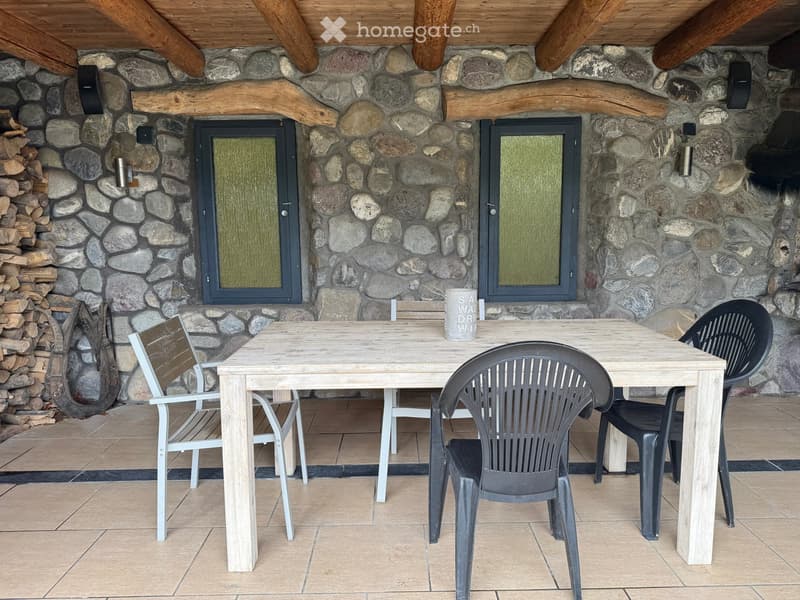 Outdoor seating area with a wooden table and four chairs, stone wall background, and a stack of firewood.