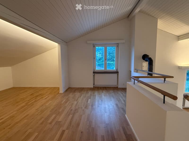Empty room with wooden flooring, white walls, and a window with a view of greenery. The room features a staircase railing on the right side and a radiator below the window.