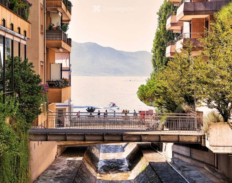 A scenic view of a canal with a bridge, surrounded by modern buildings and lush greenery, overlooking a lake with distant mountains.