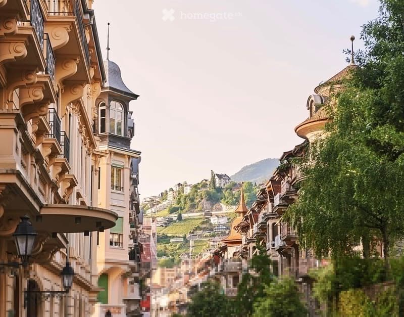 A picturesque street lined with ornate, multi-story buildings featuring balconies, decorative facades, and intricate architectural details. The background shows a hilly landscape with scattered houses and a clear sky.