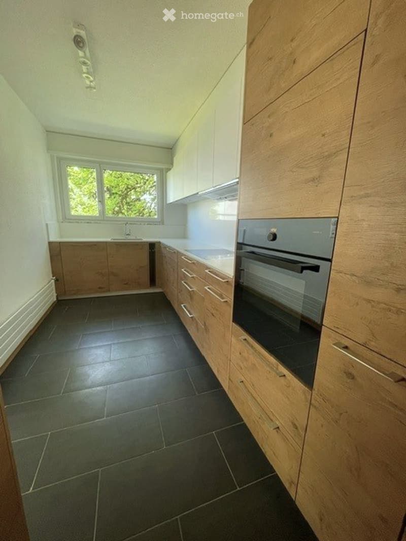 Modern kitchen with light wooden cabinets, stainless steel oven, dark tiled floor, and white countertops.