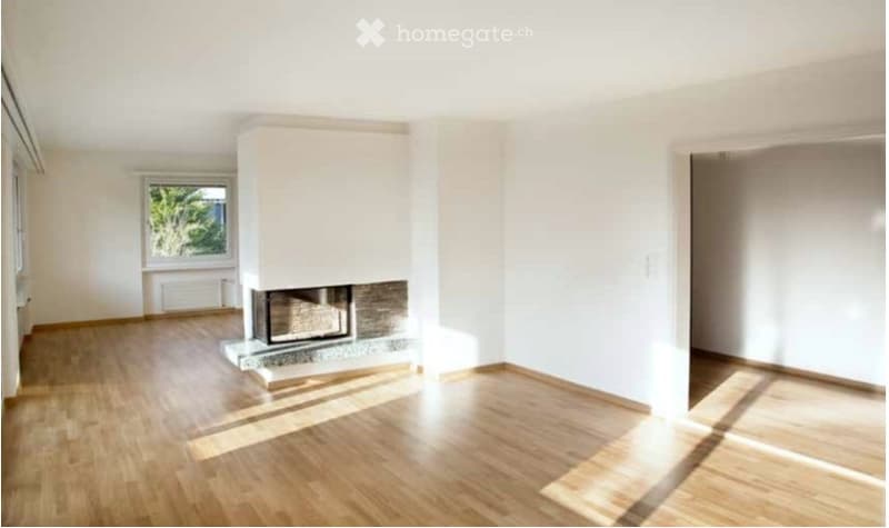 Empty room with light wooden flooring, white walls, and a central fireplace. Large window on one side allows natural light to enter.