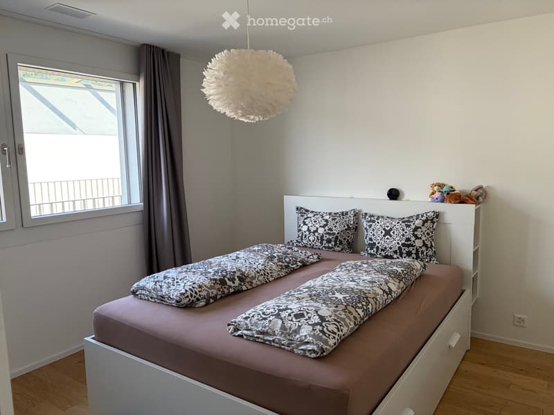 A modern bedroom with a double bed, patterned pillows, a nightstand, and a large window with a curtain. The room features a pendant light and stuffed animals on the nightstand.