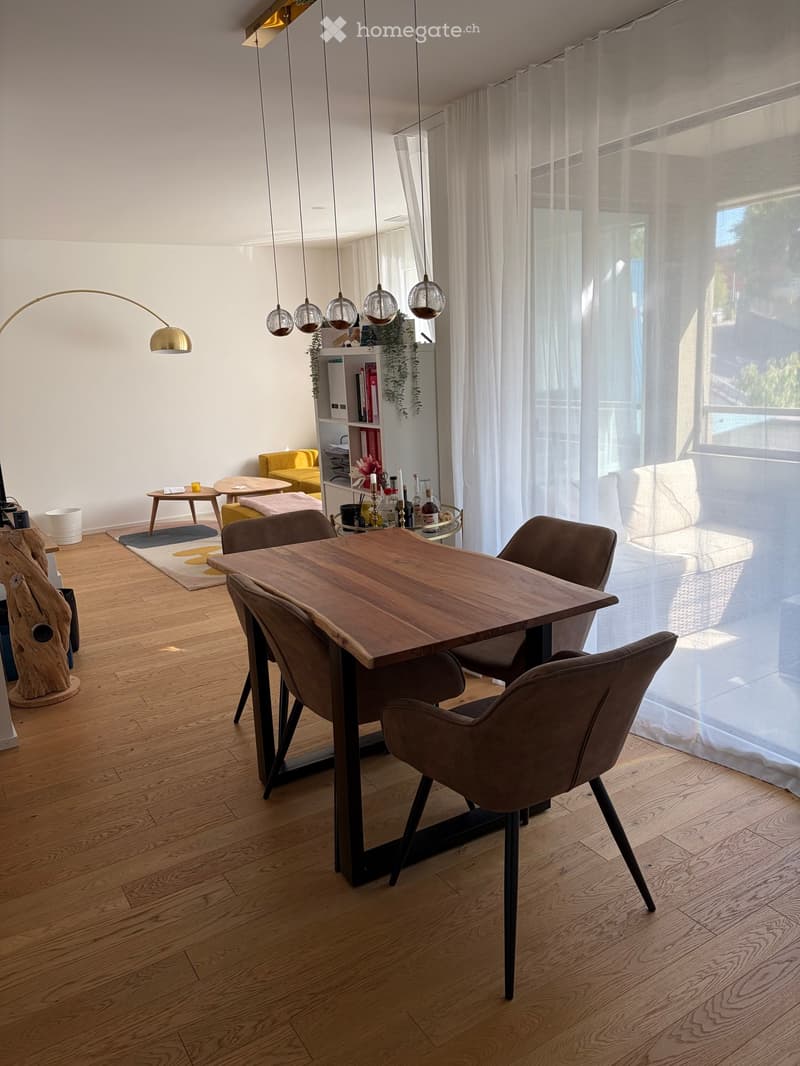 Modern dining area with wooden table, four chairs, sofa, shelf, pendant lights, and decorative items.