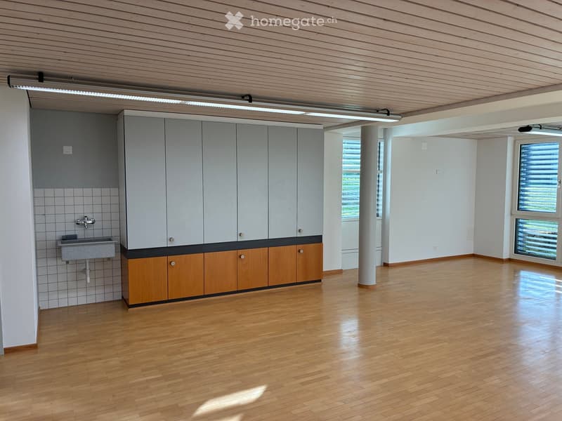 Empty room with wooden flooring, grey cabinets, sink, and large windows allowing natural light.