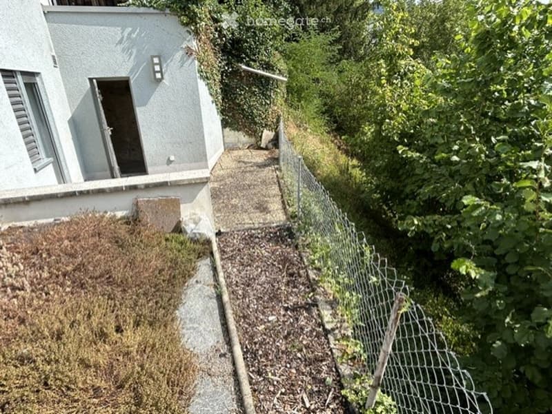 A narrow concrete pathway leading to a house entrance, bordered by a wire fence and surrounded by greenery.