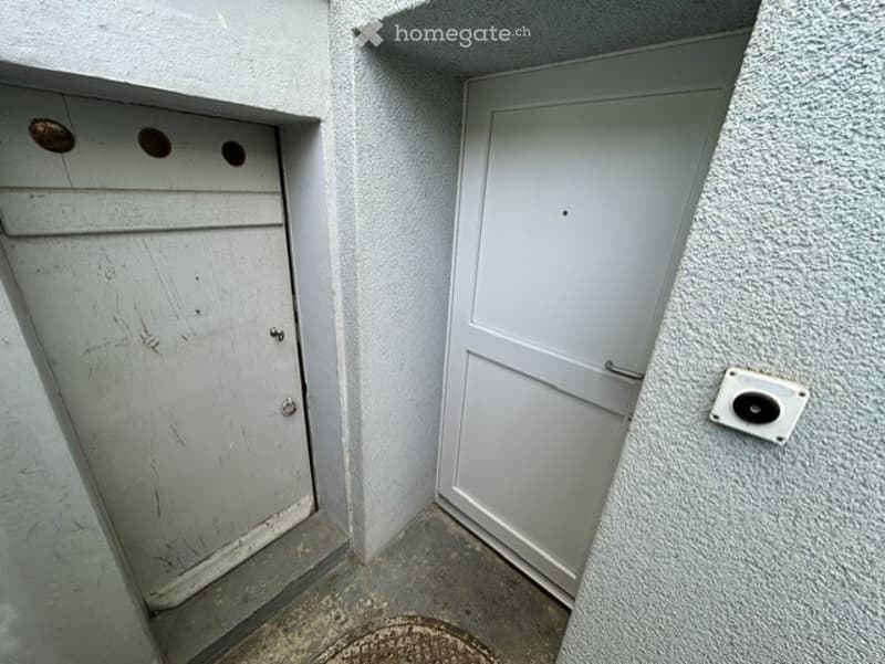 A narrow hallway with a single door on the right and a small storage compartment with three circular holes on the left wall. The floor is concrete, and the walls are painted white.