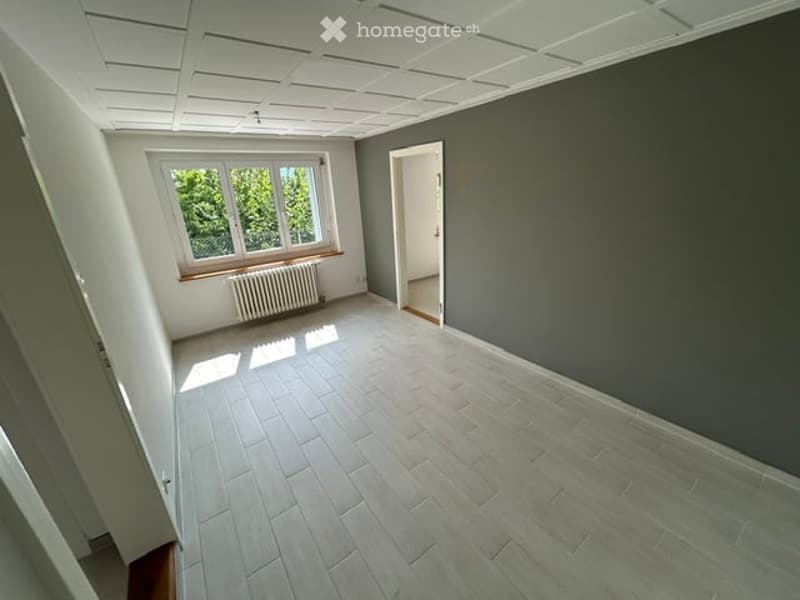 Empty room with grey walls, light grey tiled floor, large window with wooden frame, radiator below window, and an open doorway leading to another space.