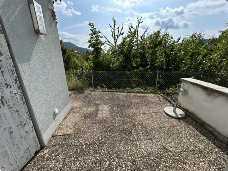 Empty concrete balcony with a view of greenery and mountains, featuring a low wall and a small stand.
