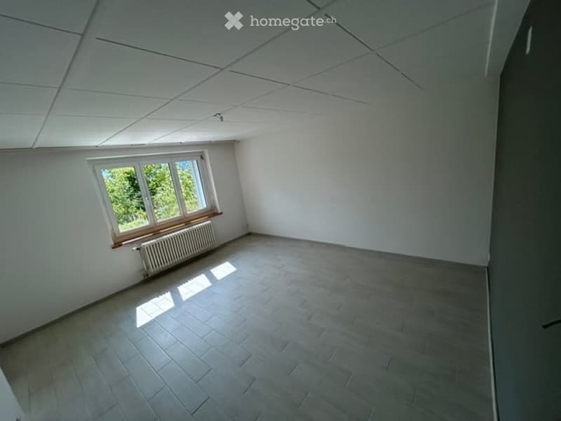Empty room with white walls, grey tiled floor, large window with wooden frame, radiator below window, and white ceiling with visible grid pattern.