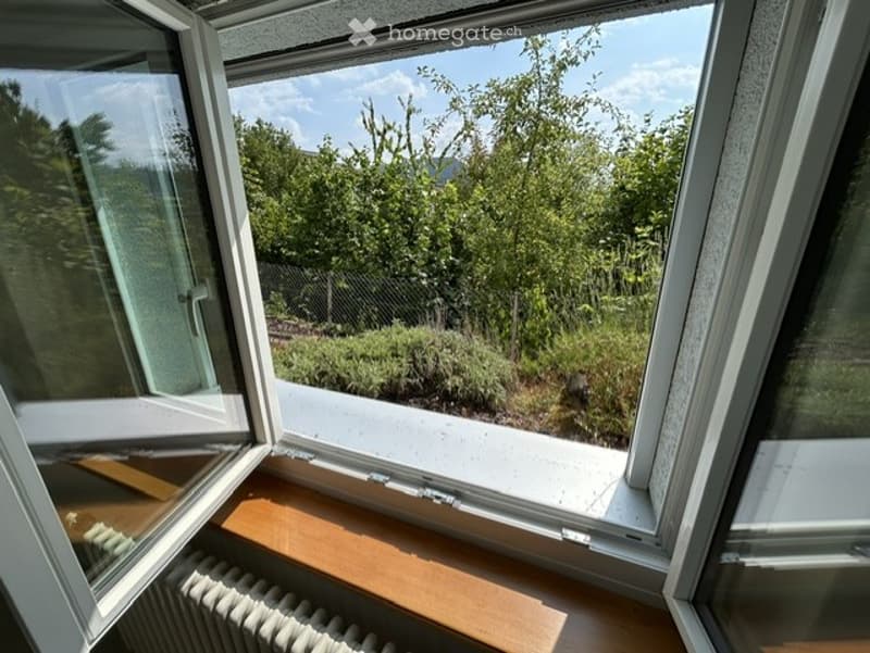 Open balcony with wooden floor, white railing, and a view of a lush green garden with trees and shrubs.