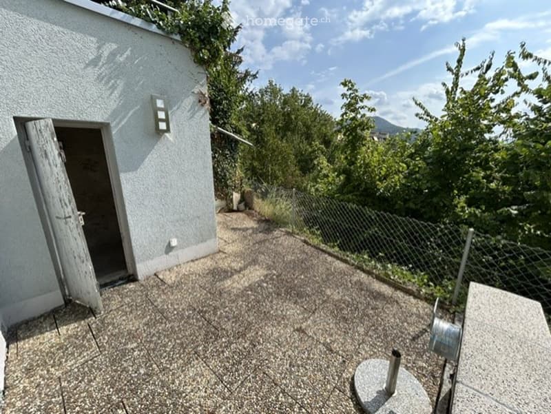 Roof terrace with concrete floor, open wooden door leading to building, metal fence, and scenic mountain view.