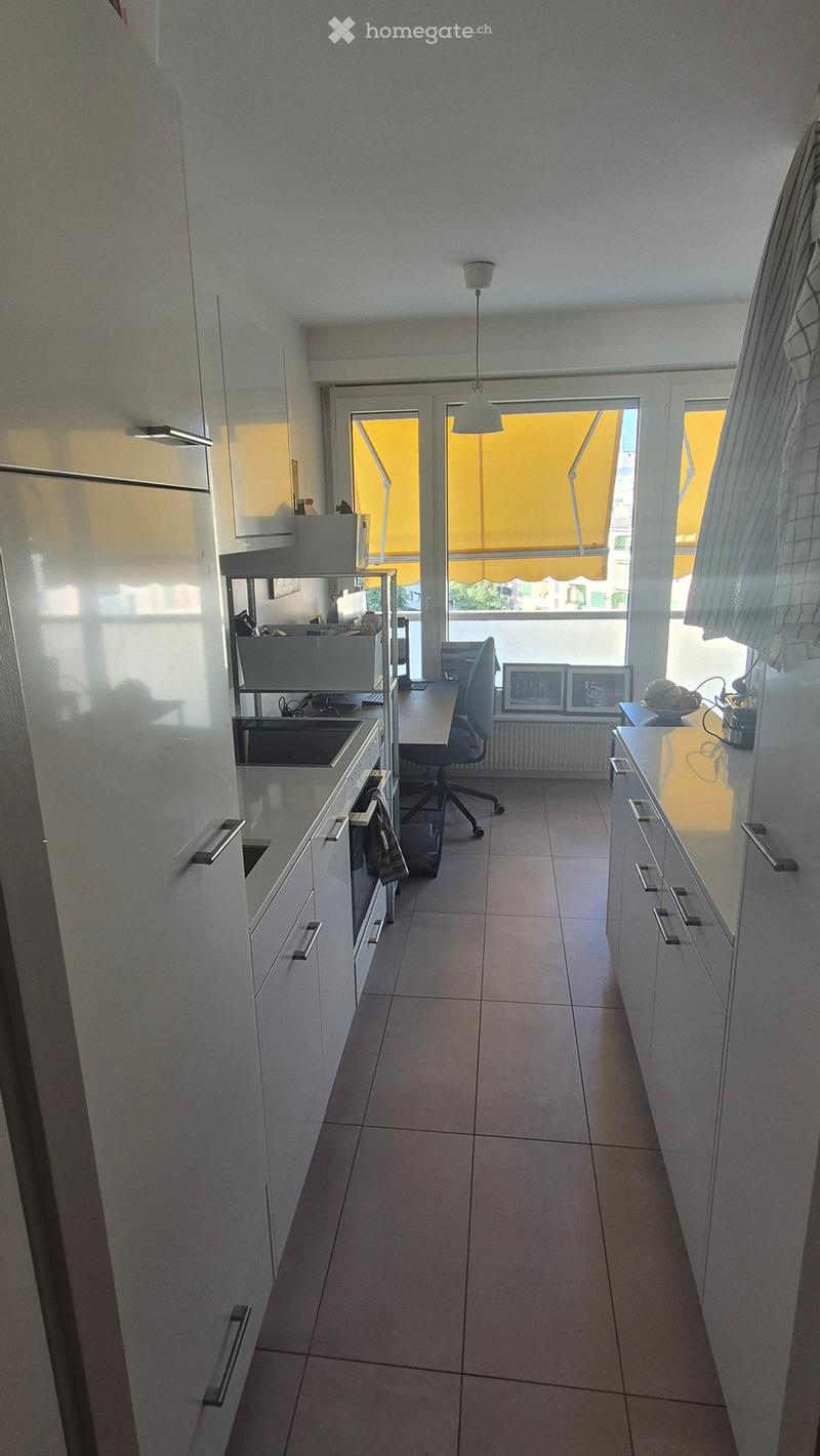 Modern kitchen with white cabinets, tiled floor, and a window with yellow blinds. Includes a sink, counter, chair, and framed pictures on the counter.