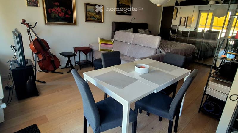 A modern living room with a white dining table, grey chairs, a sofa, a stool, a cello, and a television. The room features large windows and artwork on the walls.