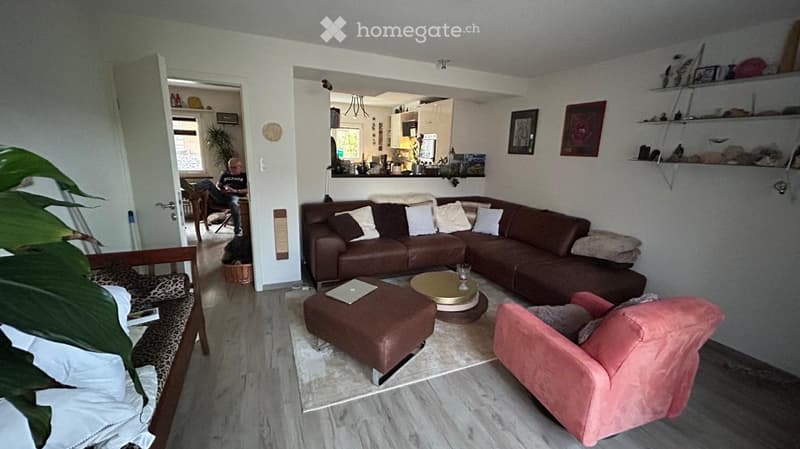 Modern living room with a brown leather sectional sofa, ottoman, coffee table, pink armchair, and shelves with decorative items. The room features wooden flooring and a view into an adjacent kitchen area.
