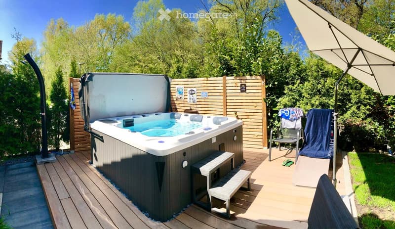 Private outdoor area with a hot tub, wooden decking, lounge chair, umbrella, and lush greenery.