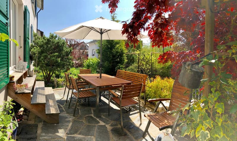 Outdoor patio with a wooden table, chairs, umbrella, and surrounding greenery.