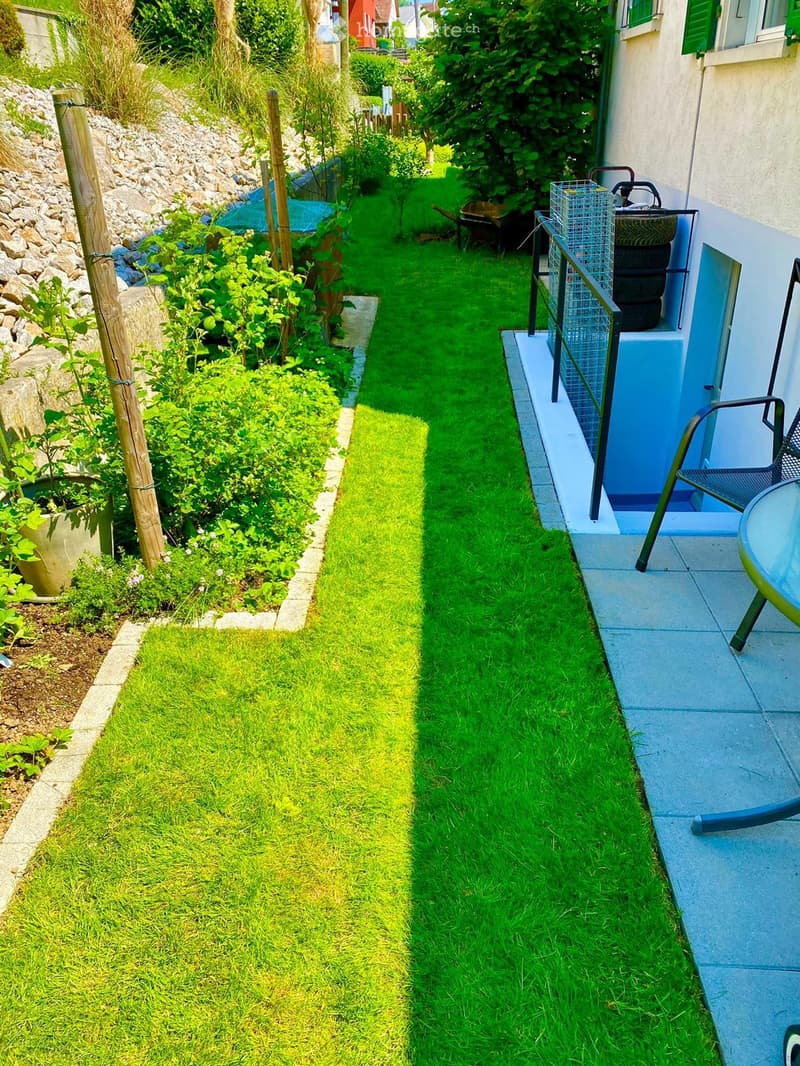 A narrow, well-maintained garden with a concrete pathway, green grass, and a small patio area with a table and chairs.