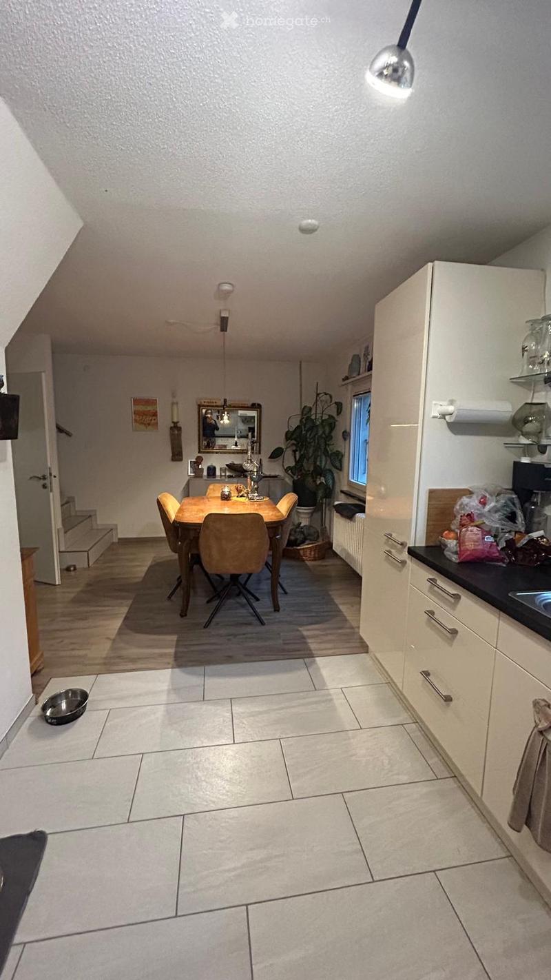 The image shows a modern kitchen and dining area with white cabinets, a black countertop, a dining table with chairs, and a large window letting in natural light. There is also a plant near the window and a dog bowl on the floor.