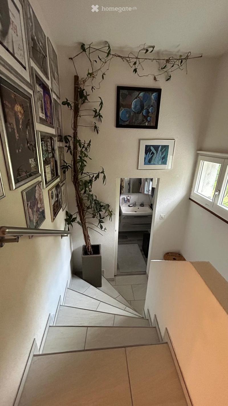 A modern hallway with tiled flooring, a glass railing, and a potted plant. Several framed artworks are displayed on the walls, and a small bathroom is visible at the end of the hallway.