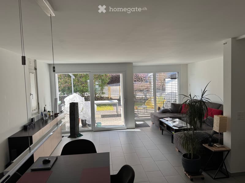 Modern living room with large sliding glass doors leading to a balcony, a sofa with red cushions, a coffee table, a potted plant, a side table with a lamp, and a workspace with a chair and laptop.