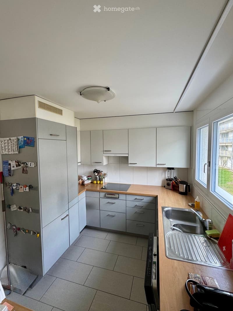 Modern kitchen with white cabinets, wooden countertops, stainless steel sink, and various appliances including a refrigerator, stove, and blender. The space is well-lit with natural light from a large window.