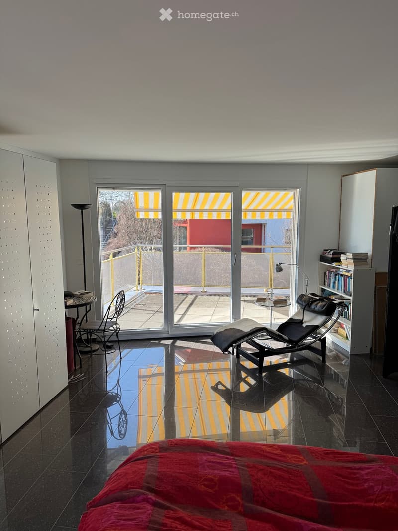 Modern bedroom with large windows leading to a balcony, featuring a red bedspread, a black leather lounge chair, a small table, and a bookshelf.
