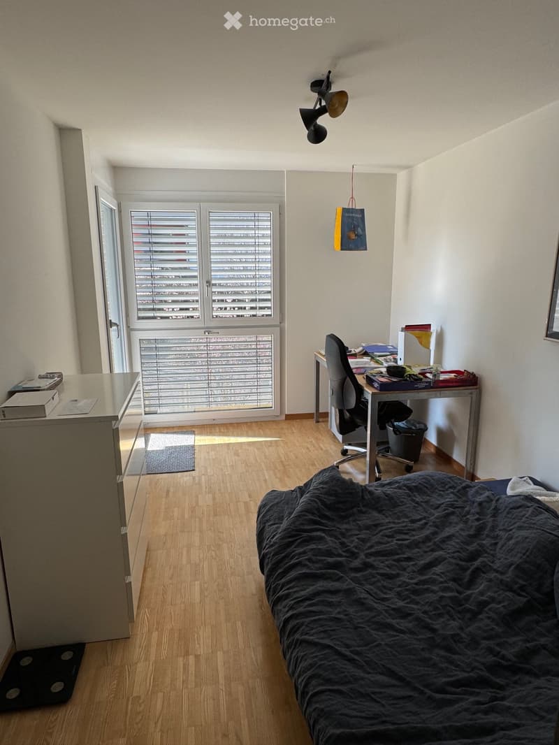 A modern bedroom with a wooden floor, a large bed with dark bedding, a desk with a chair, and a window with blinds.