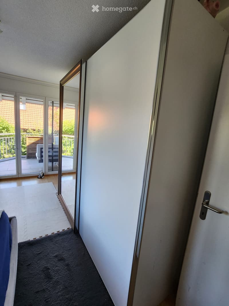 A narrow hallway with a white wall, a closed door, and an open sliding glass door leading to a balcony area with outdoor furniture visible.