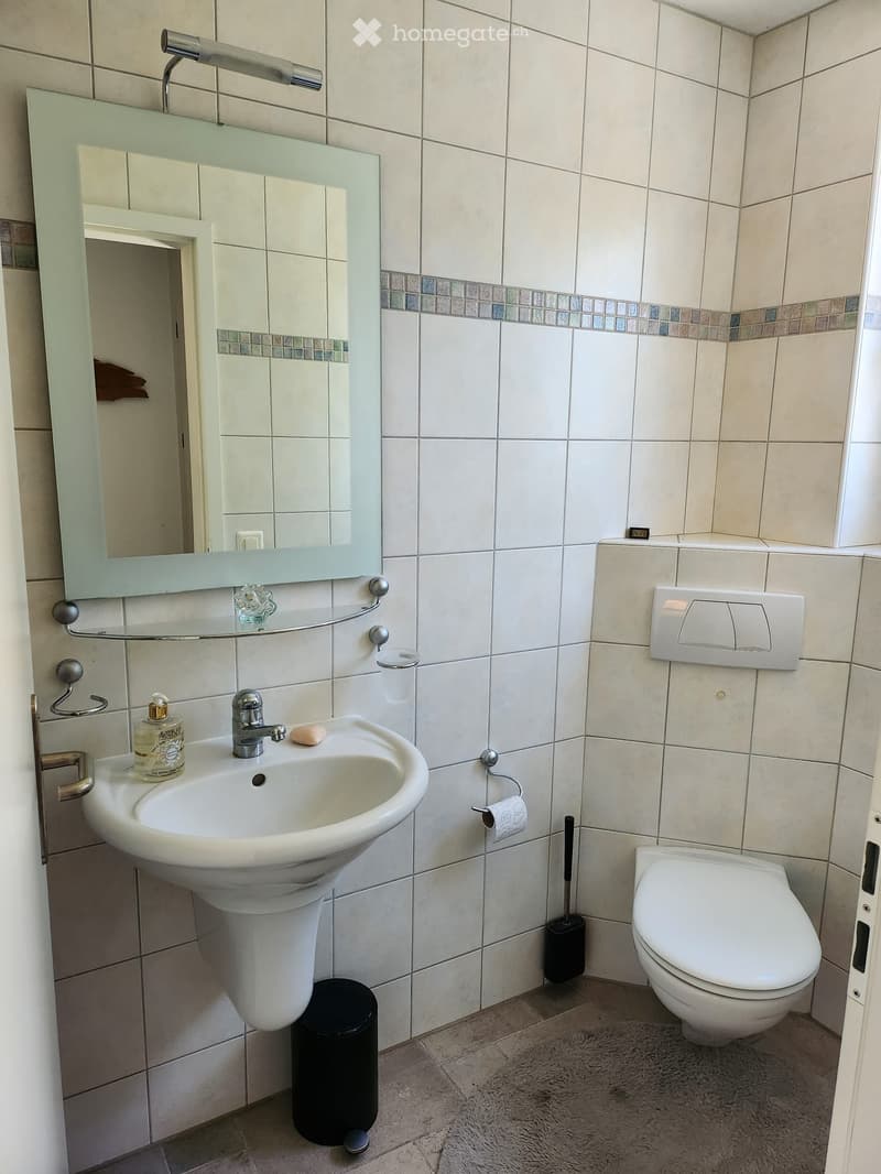 Modern bathroom with white tiles, a sink with a modern faucet, a large mirror with a built-in shelf, a toilet, and a toilet paper holder. A soap dispenser and a trash can are also visible.