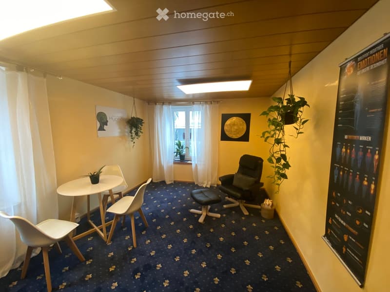 A small, cozy room with a round white table, four white chairs, a black leather sofa with a matching stool, and a potted plant on the windowsill. The walls are painted yellow, and the floor has a blue carpet with a floral pattern. There are posters on the