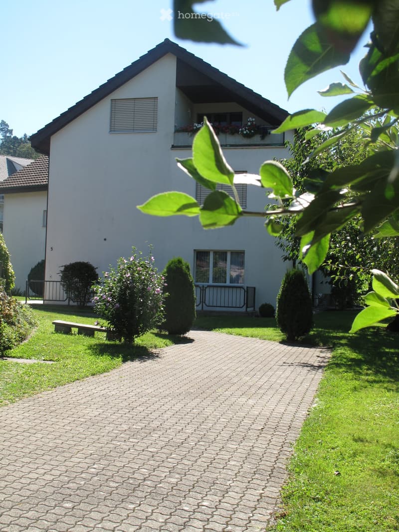 Two-story house with white exterior, dark roof, and balcony on the upper level. The front yard features a paved pathway, green grass, and trimmed shrubs.
