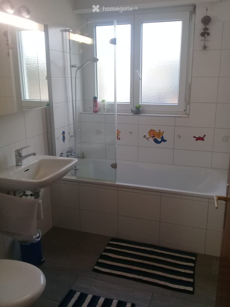 Bathroom with a sink, bathtub, showerhead, and decorative mermaid tiles on the wall. Includes a towel, rug, and small plant on the windowsill.