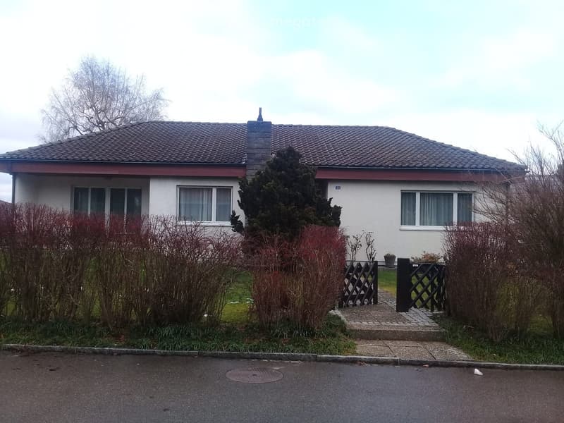 Two-story house with a tiled roof, white exterior, and multiple windows. The house is surrounded by bushes and trees, with a small garden area and a paved pathway leading to the entrance.