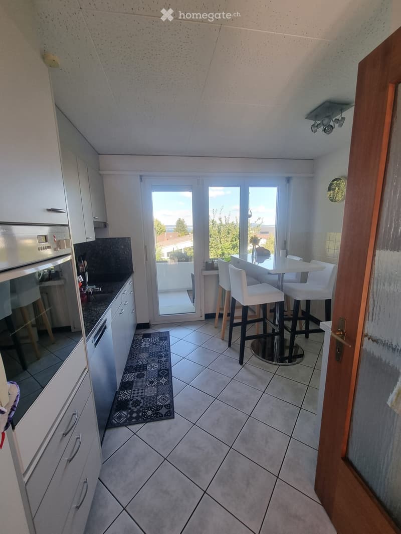 Modern kitchen with white cabinets, stainless steel oven, tiled floor, and a small dining area with white chairs and a table. A rug is placed near the kitchen island.