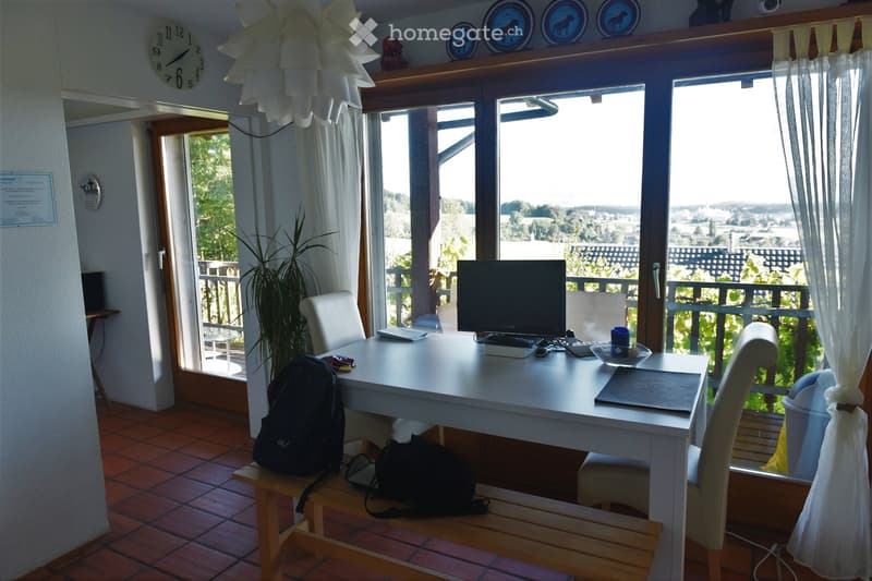Bright room with large windows offering a scenic view, furnished with a desk, chairs, a screen, and a backpack.