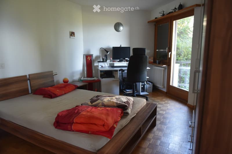 Well-lit bedroom with a wooden bed, red and beige bedding, a desk with a computer, and a large window with a view of greenery.