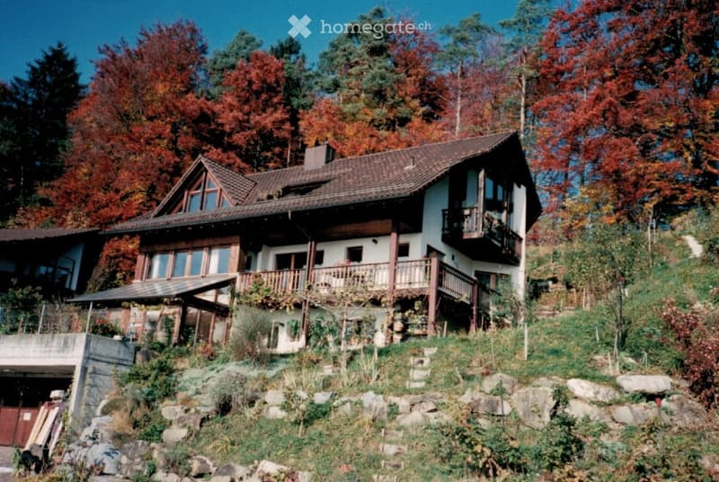 Two-story house with a sloped roof, wooden balcony, and stone foundation, surrounded by autumn foliage.