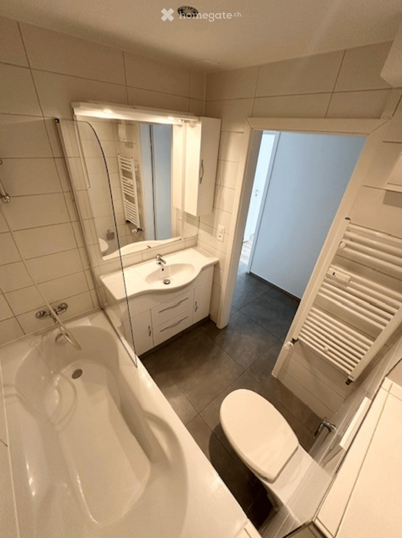 Modern bathroom with a bathtub, toilet, sink, mirror, and cabinet. The space features tiled walls and a radiator near the door.