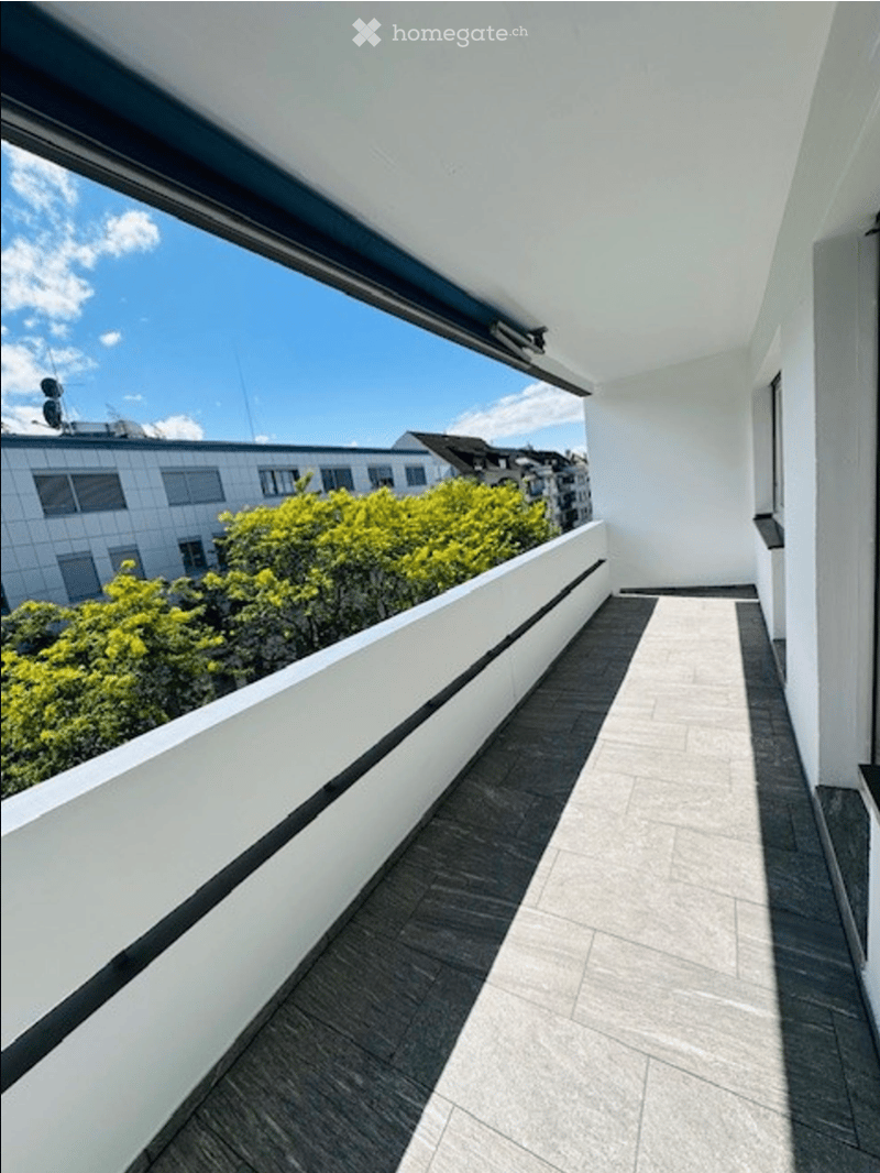 Modern balcony with white walls, tiled flooring, and a view of surrounding buildings and greenery.