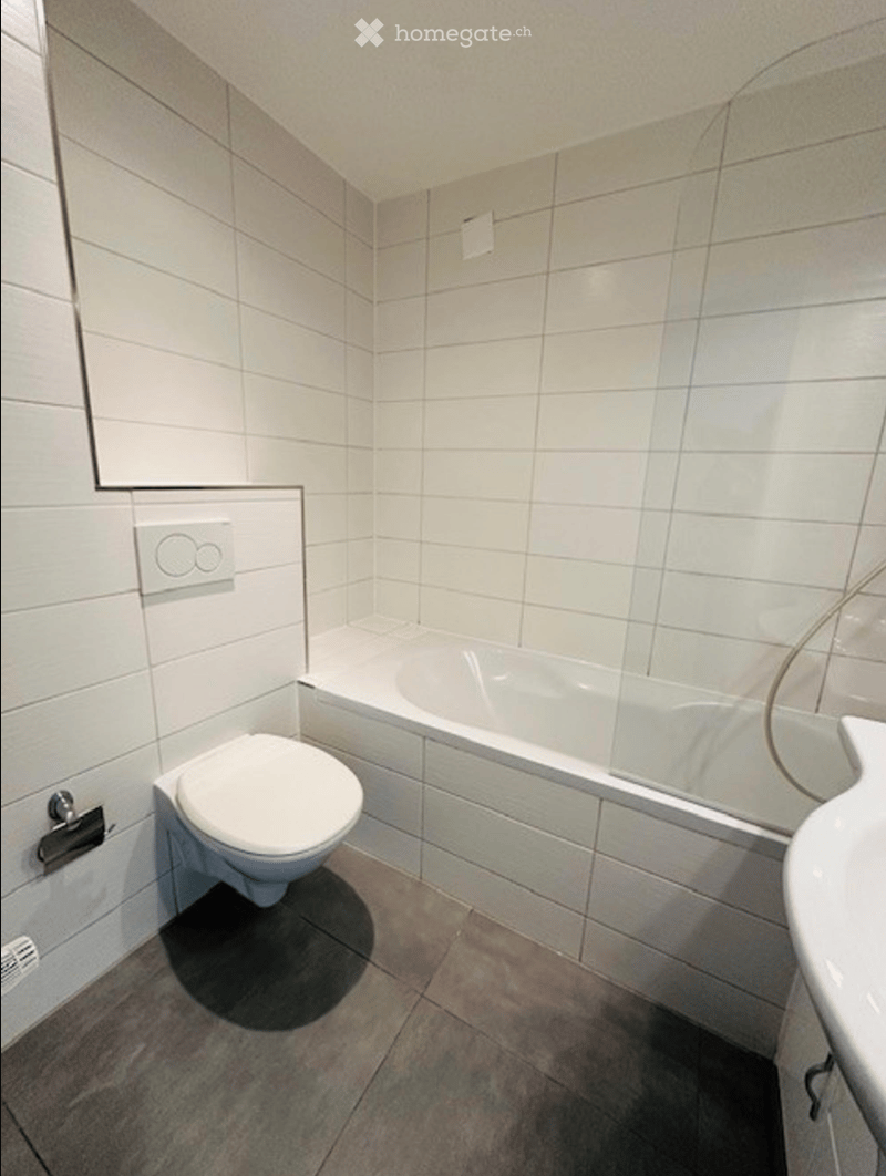 Modern bathroom with white tiled walls and floor, featuring a toilet, bathtub, and sink.