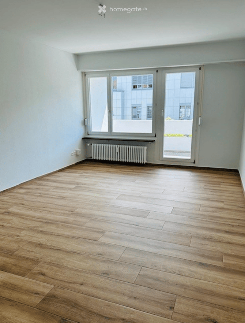 Empty room with wooden flooring, white walls, large windows, and a radiator under one window.