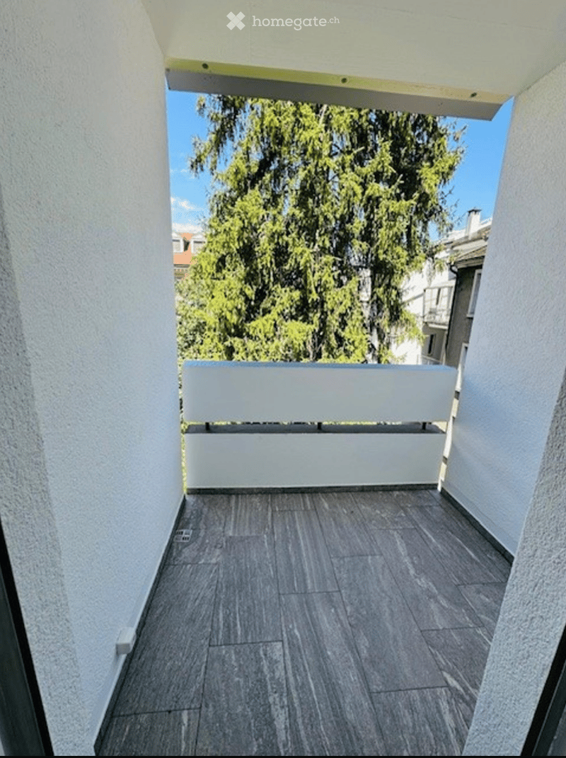 Balcony with grey tiled floor, white railing, and a view of a large tree and neighboring buildings.