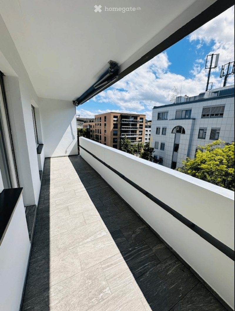 Modern balcony with white railings, dark tiled flooring, and a view of surrounding buildings and greenery.