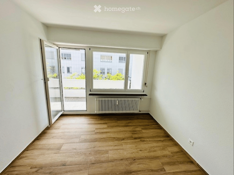 Empty room with light wooden flooring, white walls, large windows, and a radiator. No furniture or personal items present.