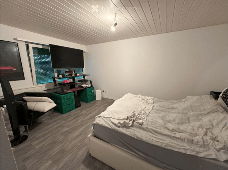 Bedroom with a bed, a chair, a desk, a screen, and a stool. The room has wooden flooring and white walls.