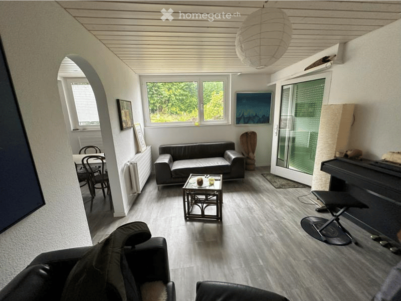 Modern living room with grey sofa, coffee table, dining area, and piano. Large windows provide natural light, and the space features a cozy seating area with additional chairs and a small table.
