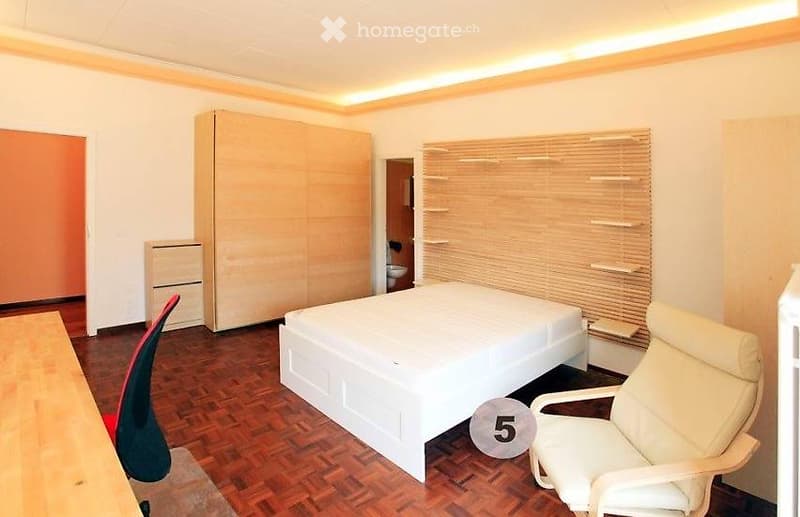 Modern bedroom with a white bed, a red and black chair, a white armchair, a large wardrobe, and a desk. The room features wooden flooring and blinds on the windows.