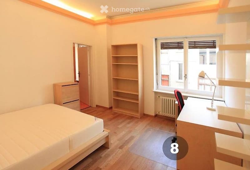 Bright bedroom with wooden flooring, a single bed, a desk with a chair, a bookshelf, and a drawer unit. Large window with blinds allows natural light.