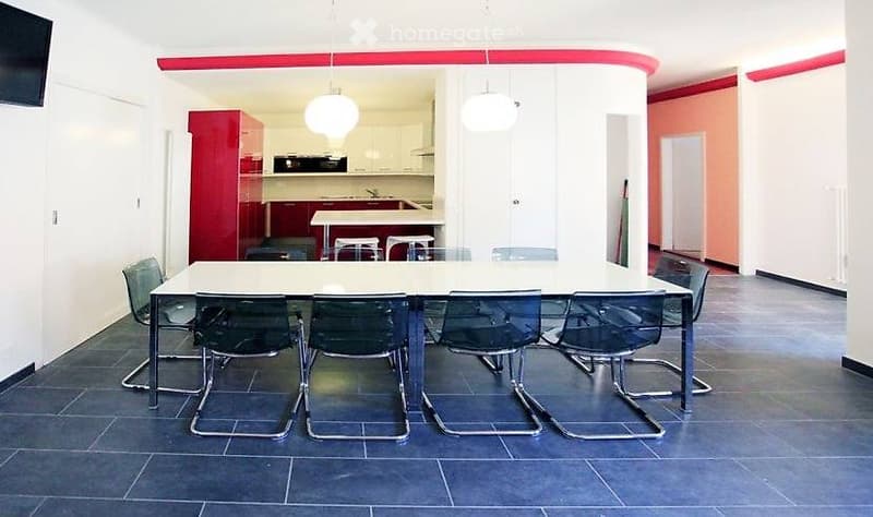 Bright, modern room with a large white table surrounded by six black chairs. In the background, a red kitchen area with cabinets, a sink, and a faucet is visible. The room features white walls, a red accent wall, and pendant lights.